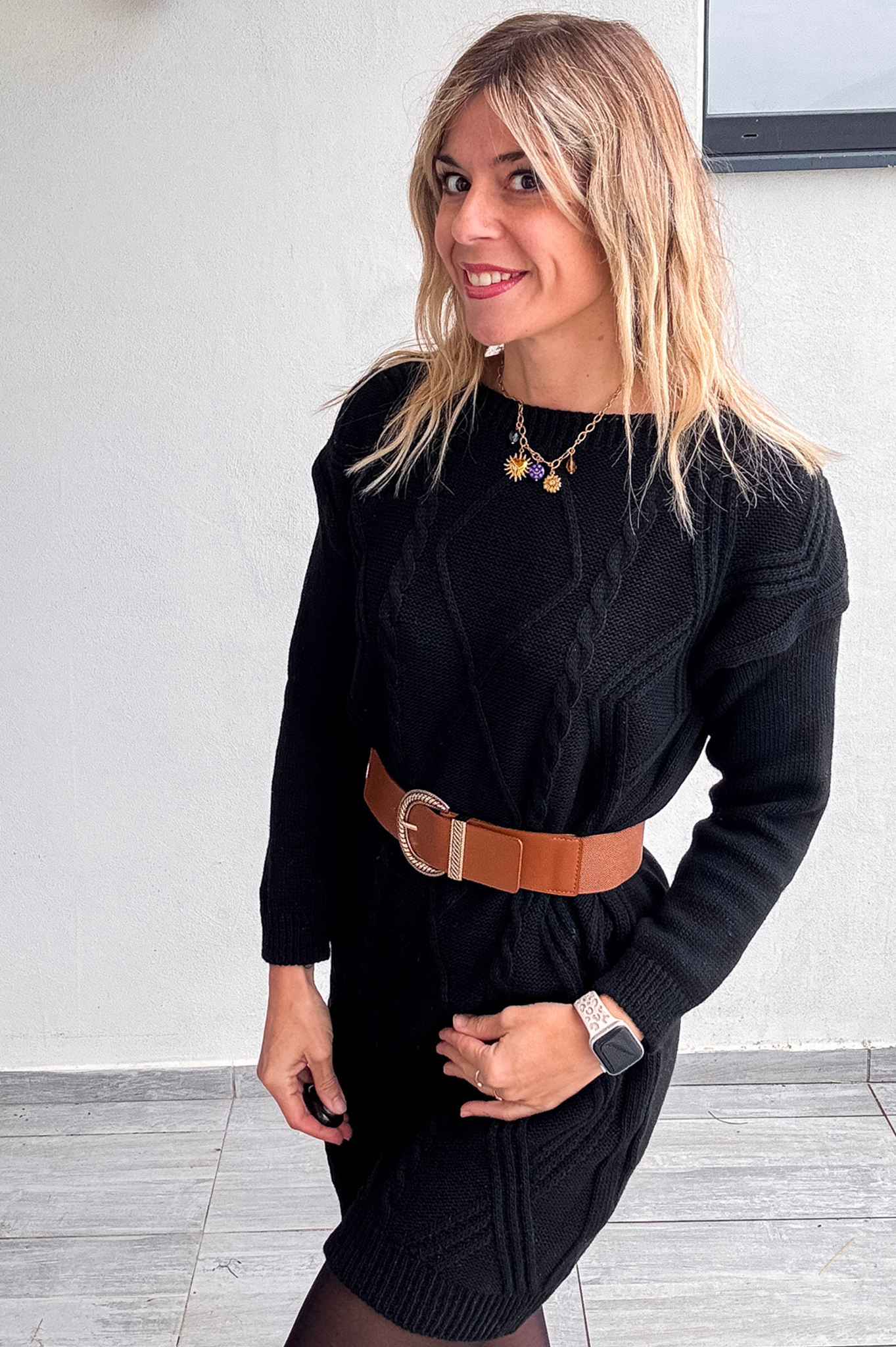 Robe pull courte noire en maille torsadée, portée avec une large ceinture marron, des collants noirs et des bottines.