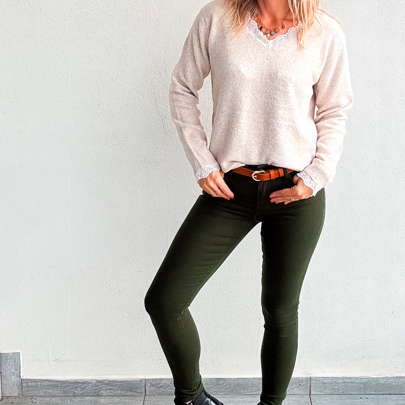 Pantalon skinny vert kaki foncé stretch, porté avec un pull beige col V à dentelle, une ceinture marron et des bottines noires.
