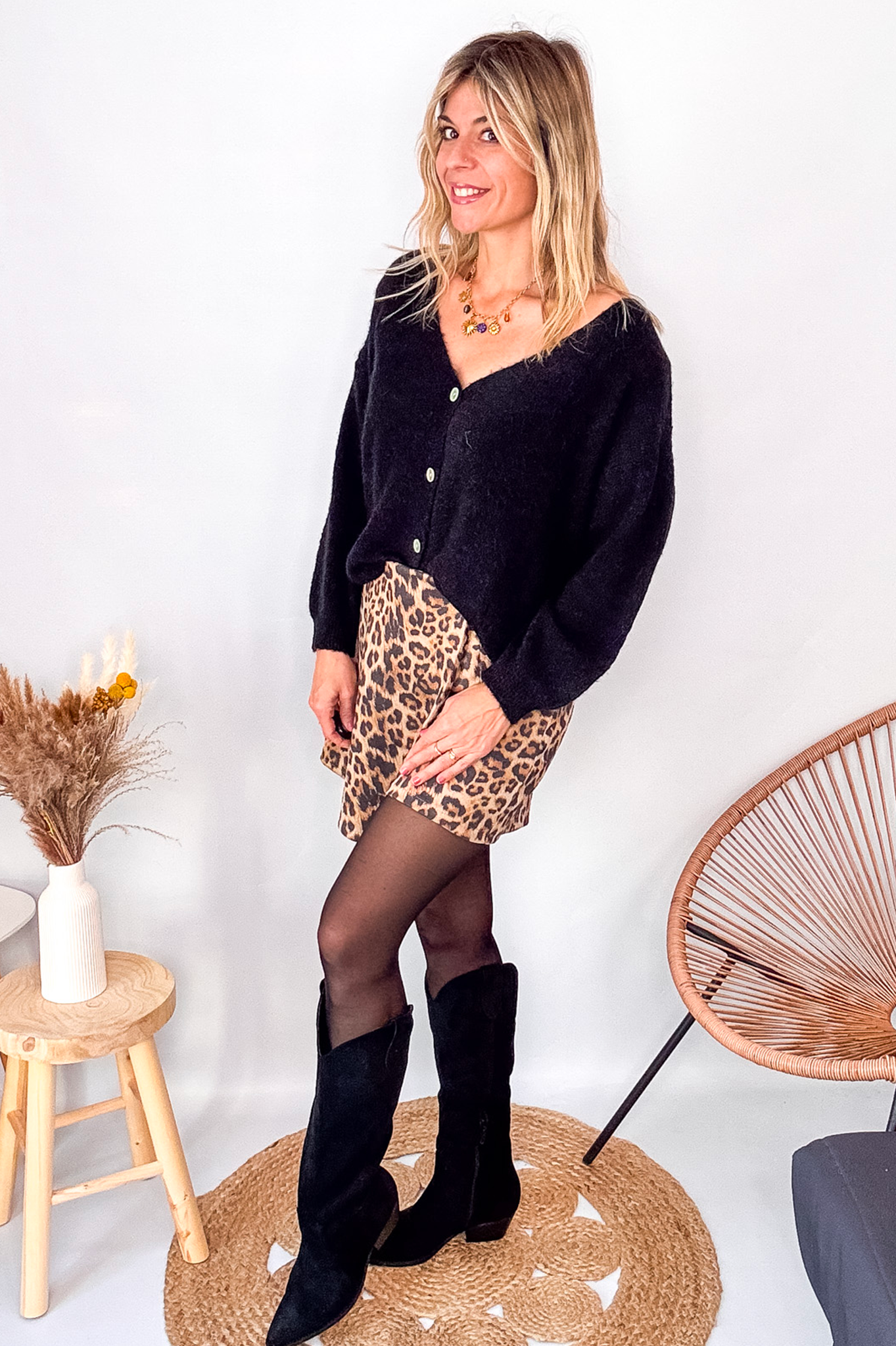 Gilet cardigan noir Baby Alpaga col V, porté sur une jupe courte imprimée léopard et des bottes noires hautes.