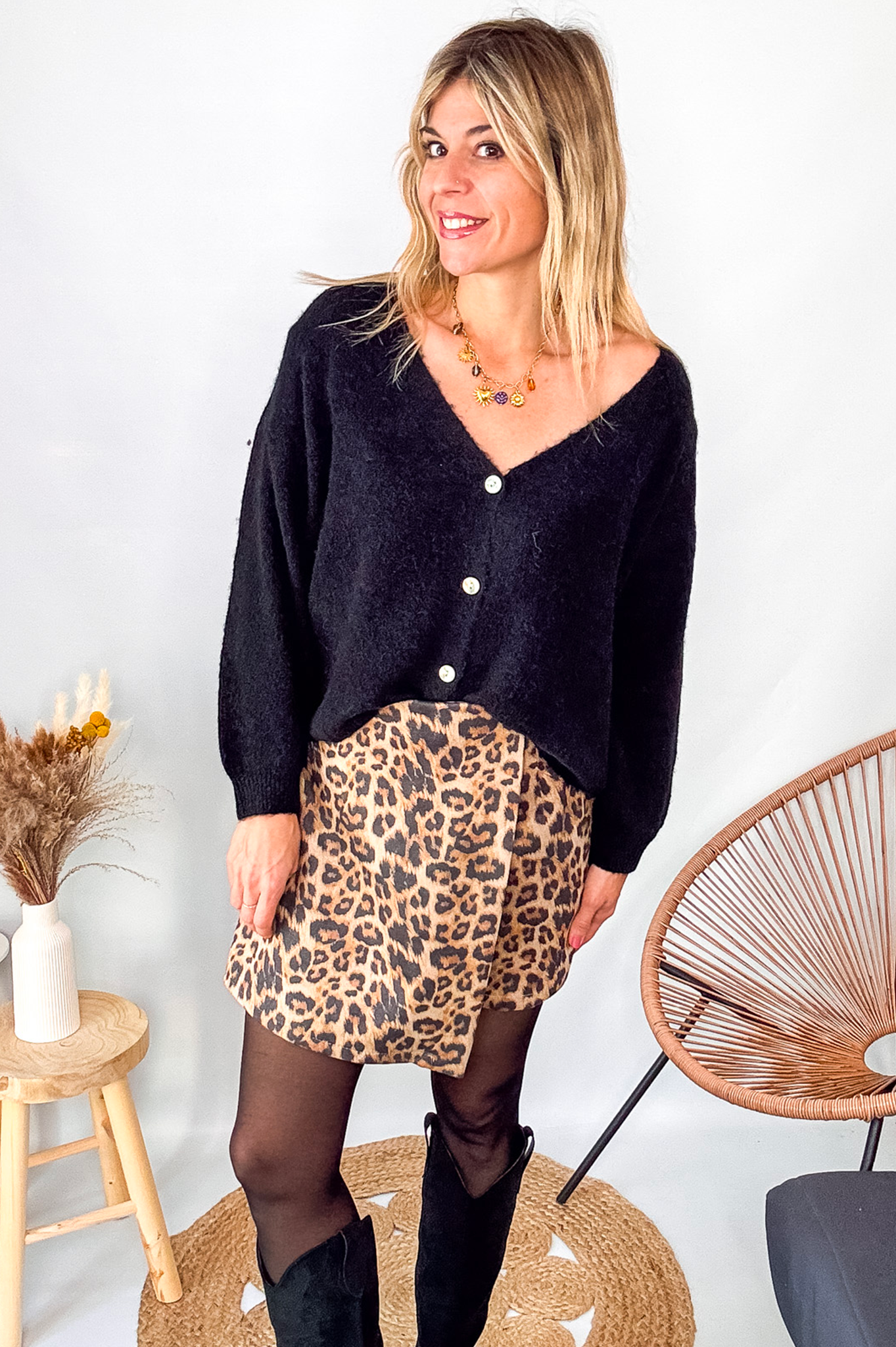 Gilet cardigan noir Baby Alpaga col V, porté sur une jupe courte imprimée léopard et des bottes noires hautes.