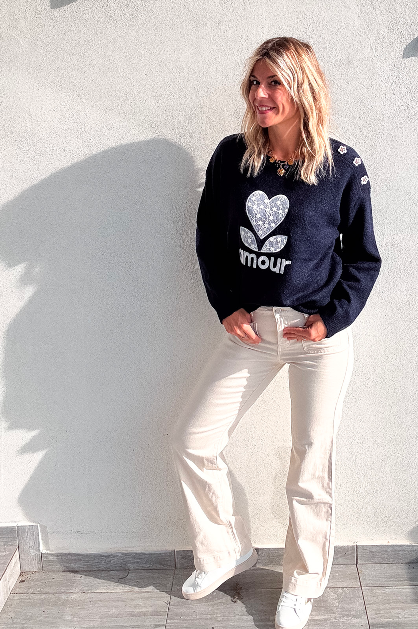 Pull bleu marine avec motif 'amour' cœur fleuri, boutons décoratifs sur l'épaule, porté avec un jean écru et des baskets blanches.