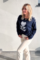 Pull bleu marine avec motif 'amour' cœur fleuri, boutons décoratifs sur l'épaule, porté avec un jean écru et des baskets blanches.