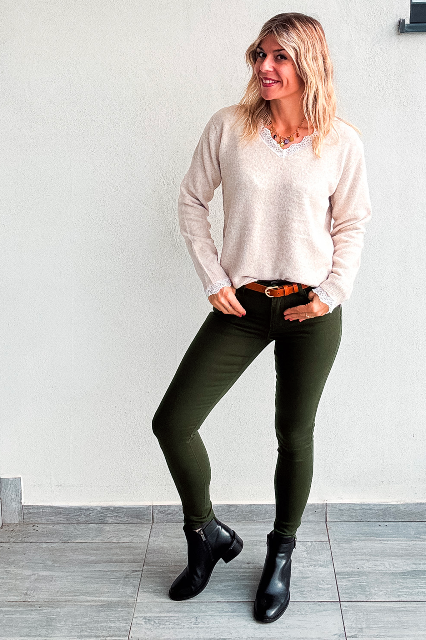 Pantalon skinny vert kaki foncé stretch, porté avec un pull beige col V à dentelle, une ceinture marron et des bottines noires.