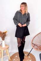 Pull gris chiné avec col en tissu imprimé carreaux, porté rentré dans une jupe noire suédine avec des collants opaques et des bottes.