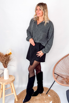 Pull gris chiné avec col en tissu imprimé carreaux, porté rentré dans une jupe noire suédine avec des collants opaques et des bottes.