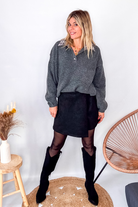 Pull gris chiné avec col en tissu imprimé carreaux, porté rentré dans une jupe noire suédine avec des collants opaques et des bottes.