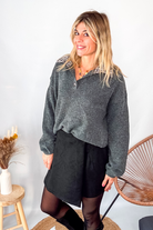 Pull gris chiné avec col en tissu imprimé carreaux, porté rentré dans une jupe noire suédine avec des collants opaques et des bottes.