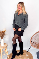 Pull gris chiné avec col en tissu imprimé carreaux, porté rentré dans une jupe noire suédine avec des collants opaques et des bottes.