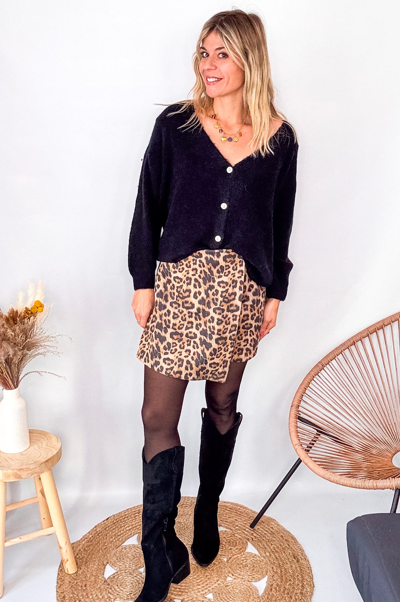Gilet cardigan noir Baby Alpaga col V, porté sur une jupe courte imprimée léopard et des bottes noires hautes.