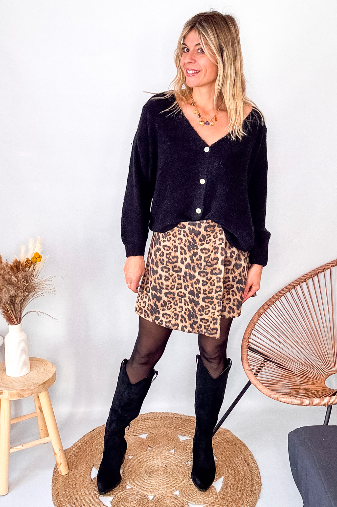 Gilet cardigan noir Baby Alpaga col V, porté sur une jupe courte imprimée léopard et des bottes noires hautes.