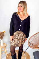 Gilet cardigan noir Baby Alpaga col V, porté sur une jupe courte imprimée léopard et des bottes noires hautes.
