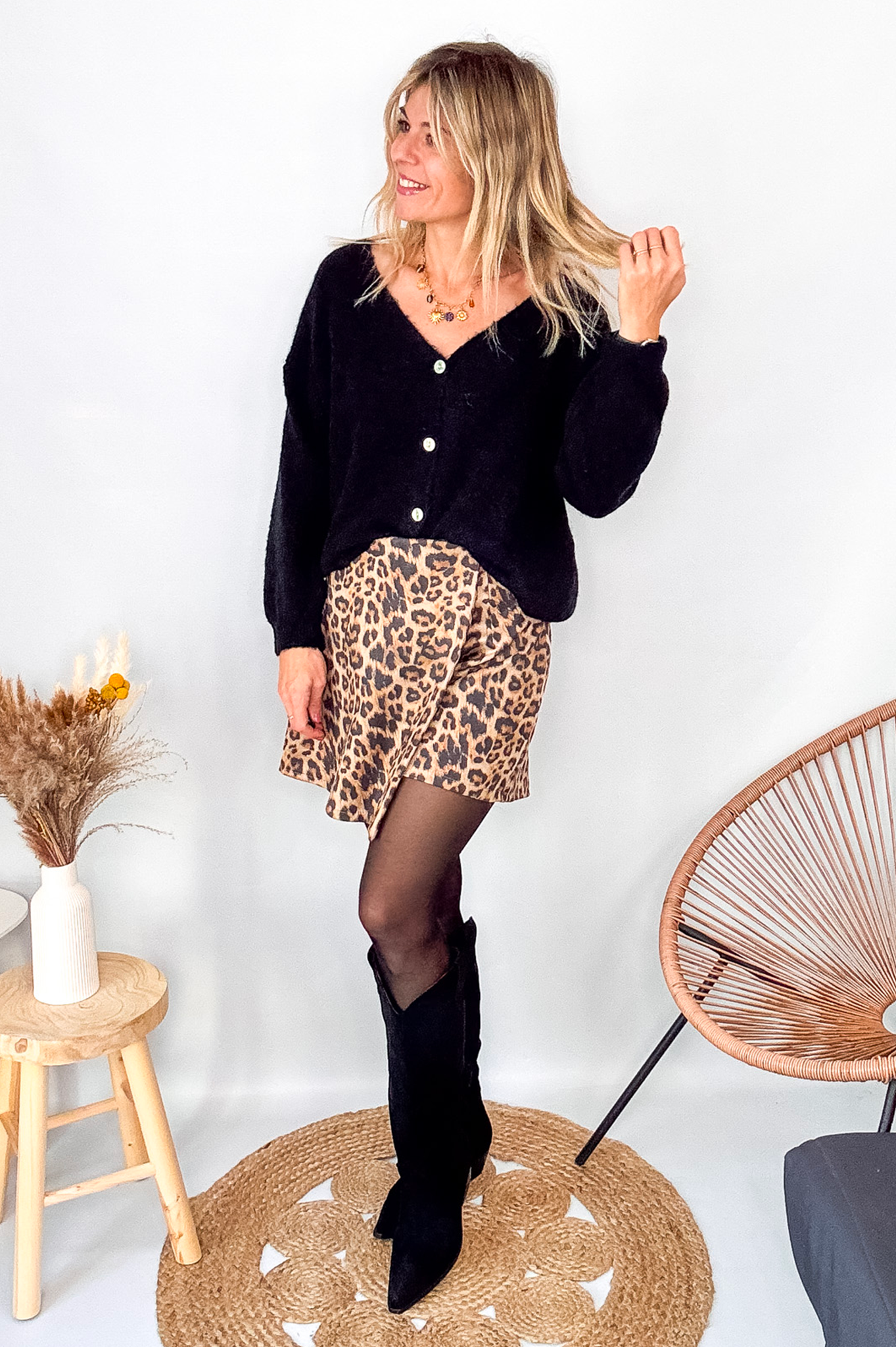 Gilet cardigan noir Baby Alpaga col V, porté sur une jupe courte imprimée léopard et des bottes noires hautes.