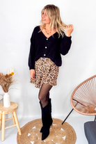 Gilet cardigan noir Baby Alpaga col V, porté sur une jupe courte imprimée léopard et des bottes noires hautes.